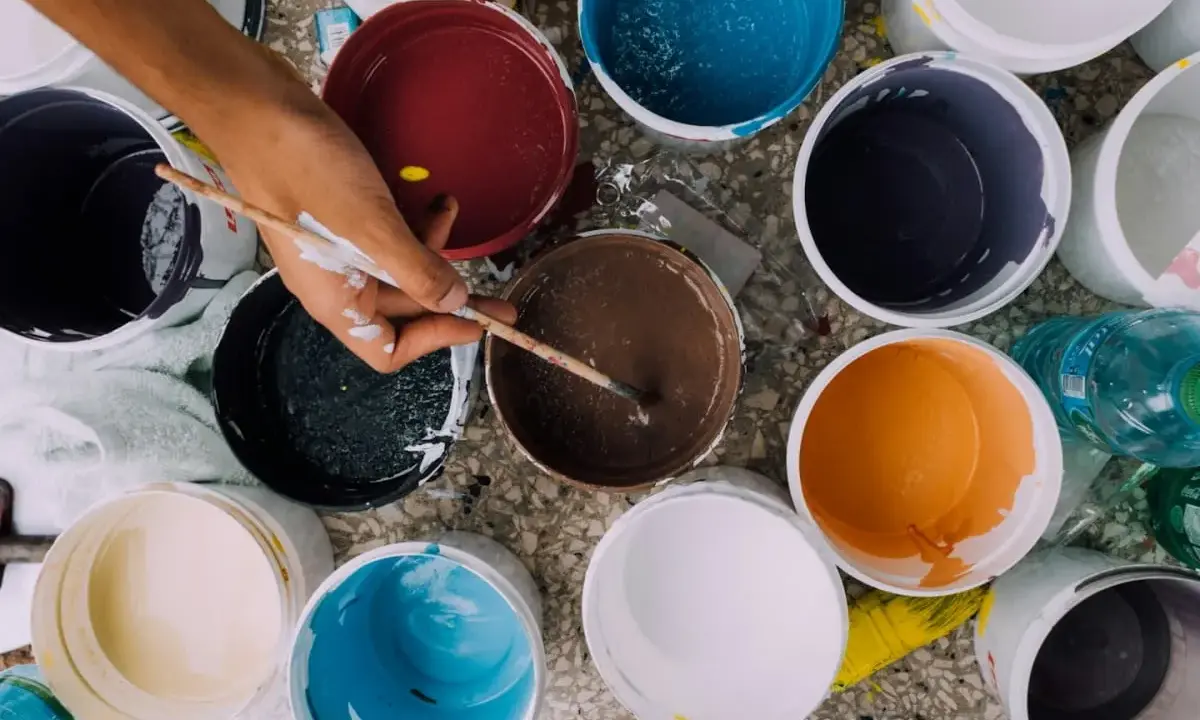 Ręka z pędzlem miesza farby w wiaderkach. Widać różne kolory, ale też potencjalne wady farby ceramicznej, jak nierównomierne rozprowadzenie.