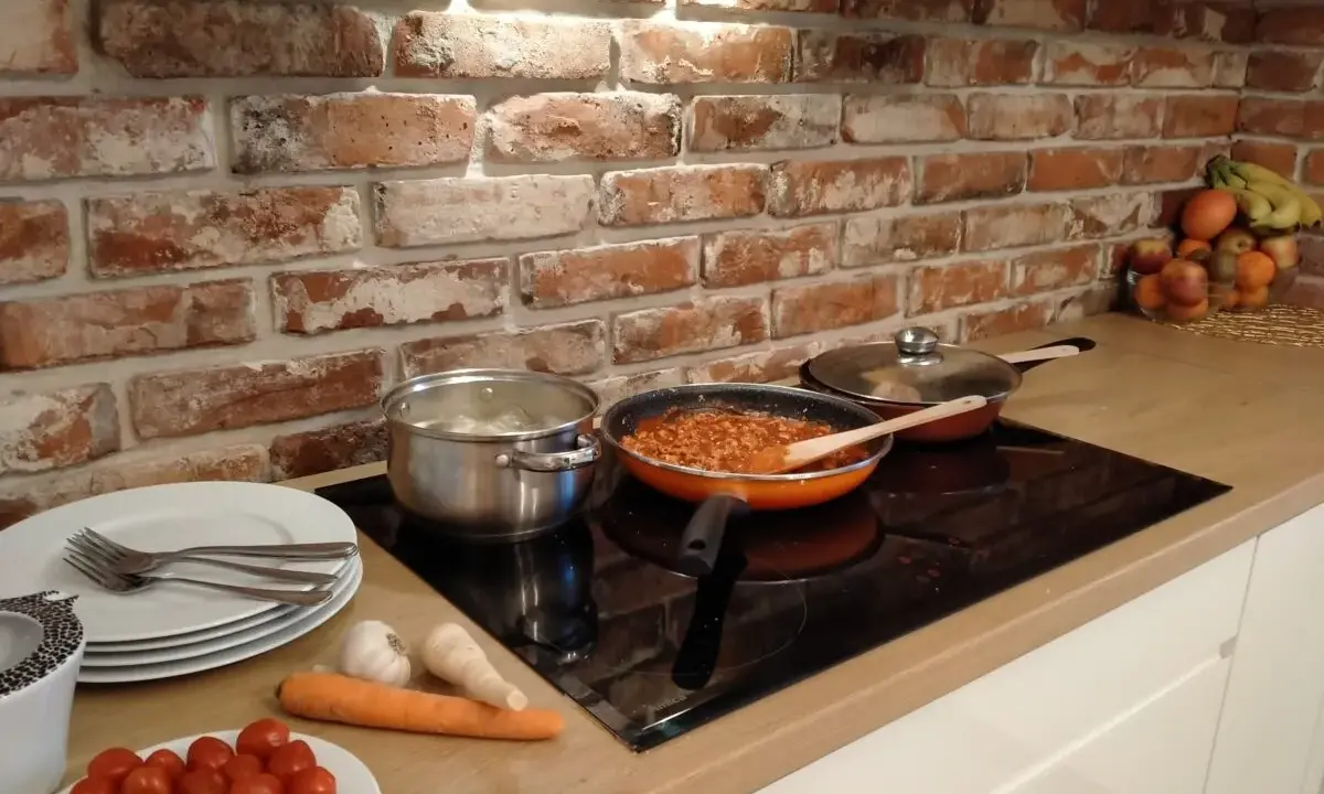 Cegła w kuchni inspiracje: gotowanie sosu bolońskiego na kuchence indukcyjnej, obok garnek z gotującymi się ziemniakami i świeże warzywa.