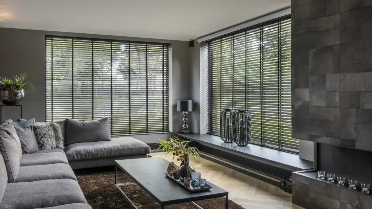 Nowoczesny salon z szarym narożnikiem i stolikiem kawowym. Duże okna zasłonięte czarnymi żaluzjami wewnętrznymi, które dodają elegancji.
