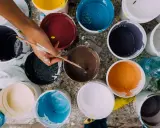 Ręka z pędzlem miesza farby w wiaderkach. Widać różne kolory, ale też potencjalne wady farby ceramicznej, jak nierównomierne rozprowadzenie.
