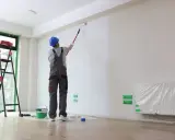 Malowanie ścian w nowym mieszkaniu. Pracownik w niebieskim kasku i szarym ogrodniczkach wałkiem pokrywa białą farbą dużą ścianę. Obok stoi drabina i wiadro z farbą. Zastanawiasz się, ile za metr malowania?