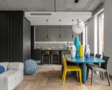 Nowoczesny, otwarty salon z jadalnią i kuchnią. Betonowy sufit, szara zabudowa kuchenna, niebieski stół i kolorowe wazony.