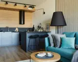Nowoczesny salon z aneksem kuchennym 22m2. Turkusowa kanapa, czarne hokery przy wyspie, drewniane akcenty i stylowe oświetlenie tworzą przytulną przestrzeń.