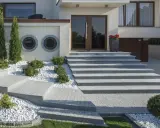Nowoczesne schody zewnętrzne z beton dekoracyjny, otoczone białym żwirem i zielenią, prowadzące do drzwi wejściowych.