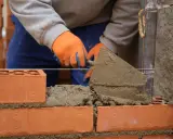 Budowa prostego grilla z cegły. Ręce w pomarańczowych rękawiczkach nakładają zaprawę kielnią na kolejne cegły.