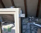 Drewniane okno z metalowym kątownikiem, gotowe do obróbki po montażu. W tle widać materiały budowlane i drewniane belki poddasza.