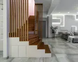 Nowoczesna, wysoka klatka schodowa aranżacje z drewnianymi stopniami i ażurową balustradą, wkomponowana w minimalistyczne wnętrze z szarą sofą i geometryczną ścianą.