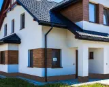 Nowoczesny dom z białym tynkiem i drewnianymi akcentami. Z czego zrobić cokół wokół domu? Widać tu kamyczki, które nadają mu ciekawy wygląd.