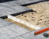 Budowa tarasu z płyt betonowych w toku. Widać układane płyty, podsypkę piaskową i obrzeża. Narzędzia budowlane leżą obok.