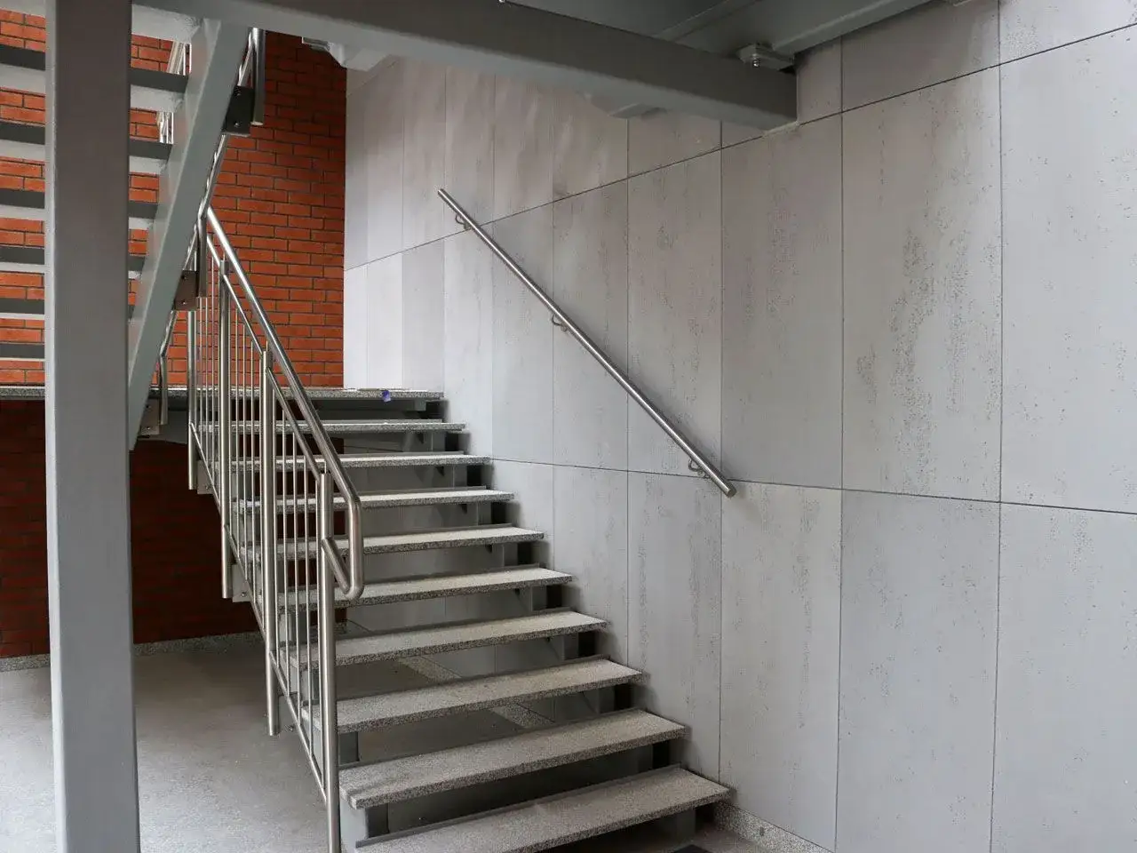 Nowoczesne schody zewnętrzne z kamiennymi stopniami i metalową balustradą, obok ściany z dekoracyjnego betonu.