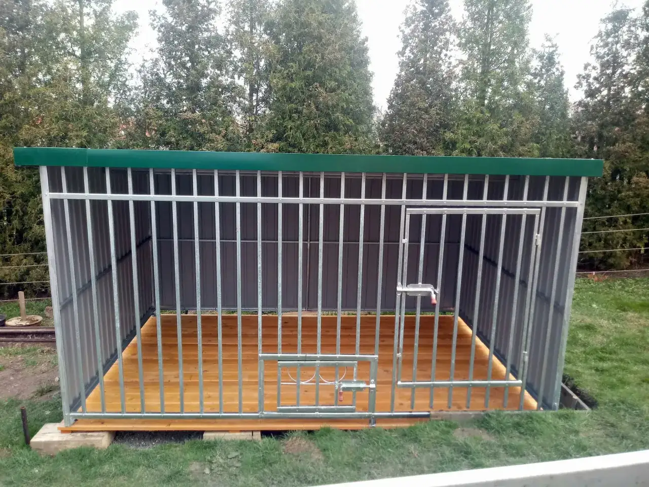 Nowy, solidny kojec dla psa z paneli ogrodzeniowych, z drewnianą podłogą i zielonym dachem, gotowy na przyjęcie czworonożnego przyjaciela.
