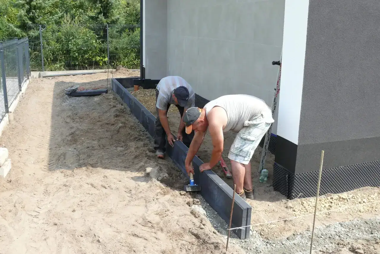Dwóch mężczyzn układa obrzeża pod kostkę. Przygotowują teren, używając narzędzi do wyrównania podłoża. Suchy beton pod kostkę proporcje to klucz do trwałej nawierzchni.