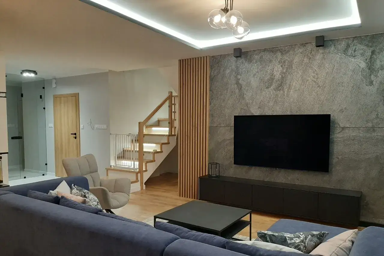 Nowoczesne lamele w salonie aranżacje: drewniana ściana z telewizorem, schody z oświetleniem, wygodna sofa i fotel.