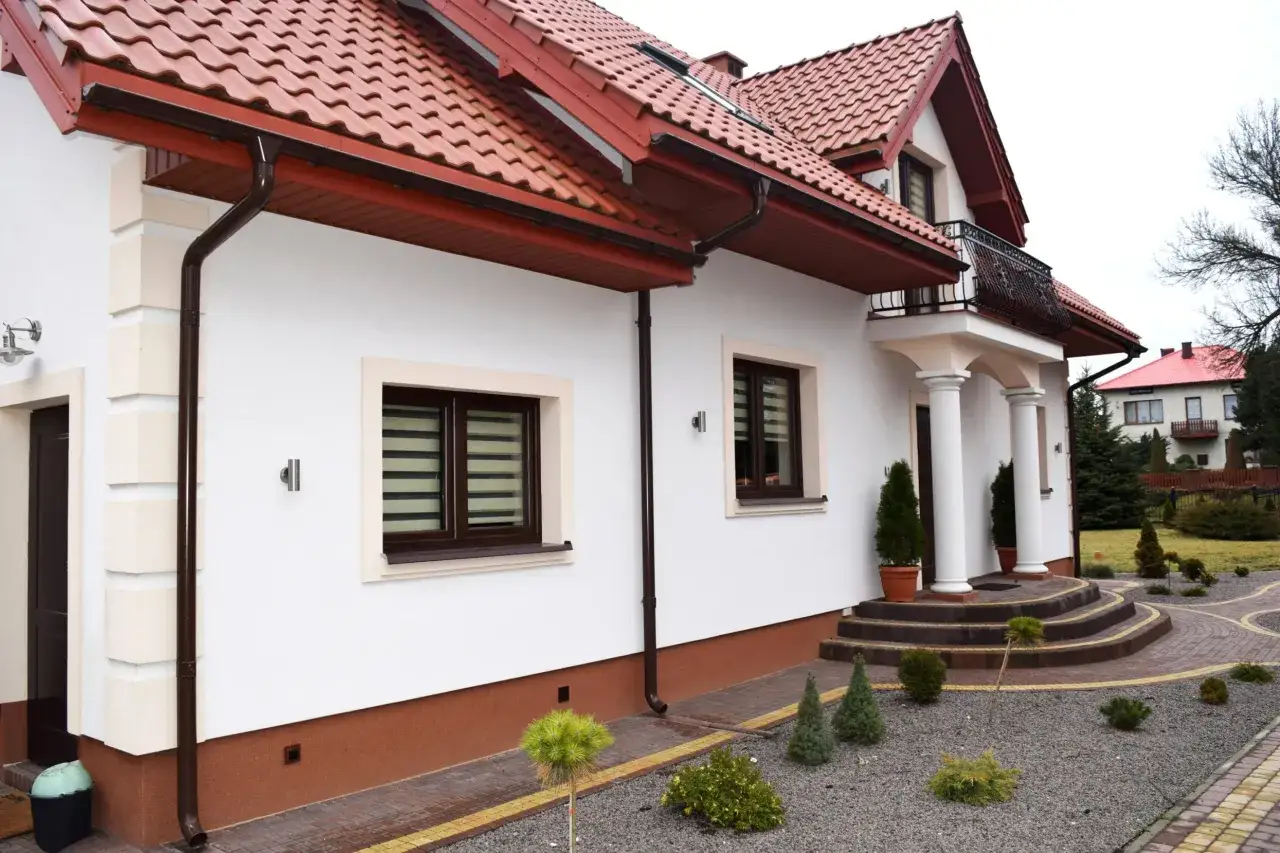 Z czego zrobić cokół wokół domu? Na zdjęciu biały dom z brązowym cokołem, czerwonym dachem i kamienną ścieżką.