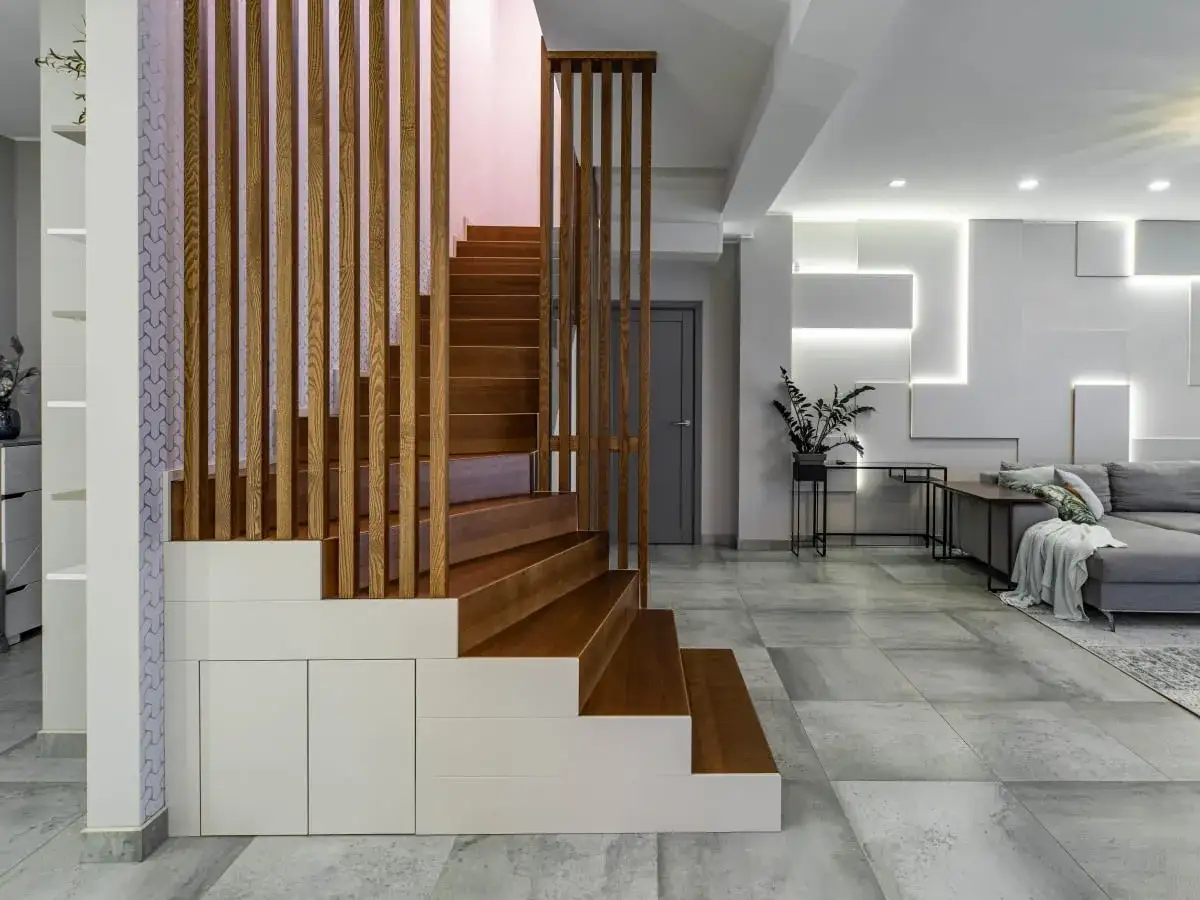 Nowoczesna, wysoka klatka schodowa aranżacje z drewnianymi stopniami i ażurową balustradą, wkomponowana w minimalistyczne wnętrze z szarą sofą i geometryczną ścianą.
