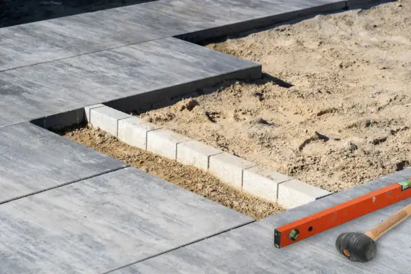 Budowa tarasu z płyt betonowych w toku. Widać układane płyty, podsypkę piaskową i obrzeża. Narzędzia budowlane leżą obok.